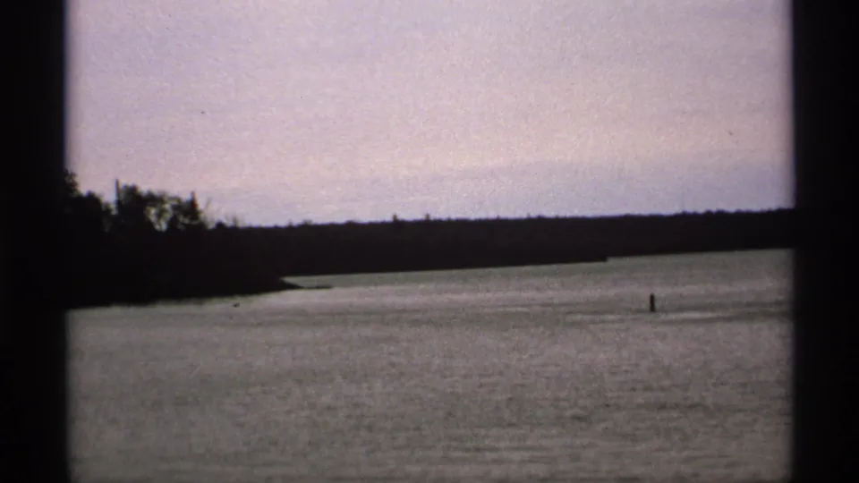 1971 Isle Royale frame showing a broad lake passage, wooded shoreline, pale sky, and a small distant buoy or marker.