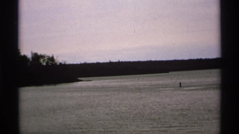 1971 Isle Royale home-movie view of calm water and a dark tree-lined shoreline, framed by visible 8mm gate edges.