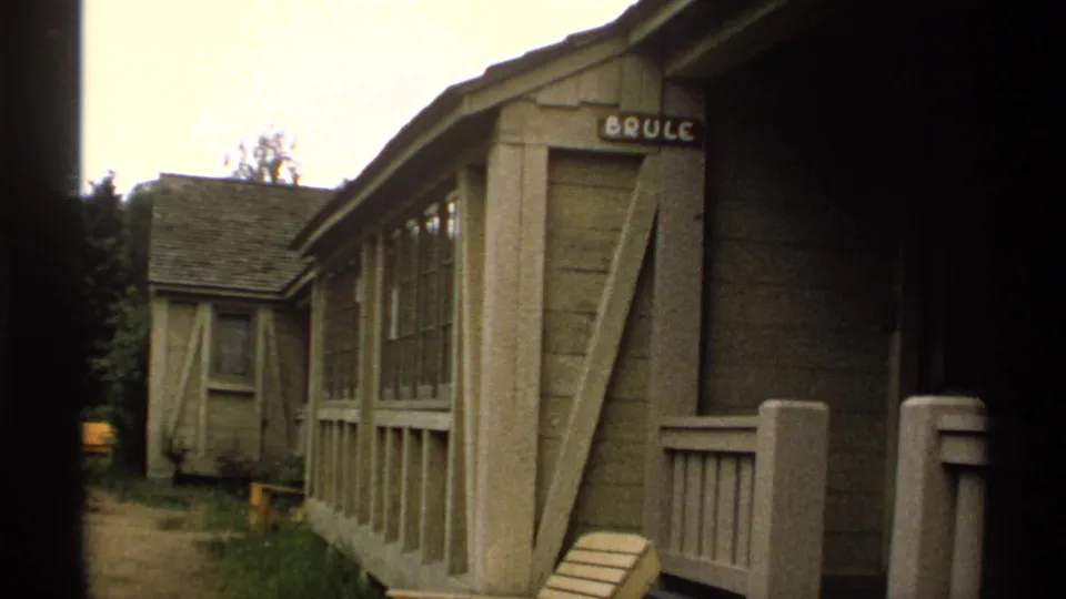 Wider 1971 Isle Royale view of a rustic wooden cabin with porch rail, windows, path, trees, and the BRULE sign above the entry.