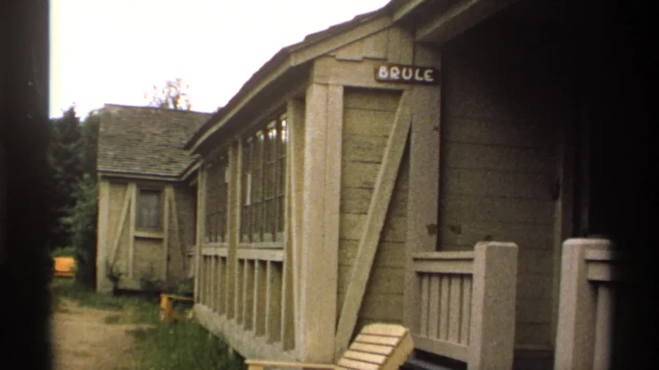 1971 Isle Royale 8mm frame of the BRULE cabin exterior, showing weathered siding, side windows, porch rail, and a wooded path.