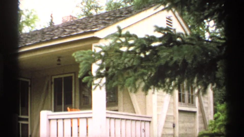 1971 Isle Royale 8mm frame showing another rustic cabin porch partly obscured by evergreen branches, with windows and pale porch railings.