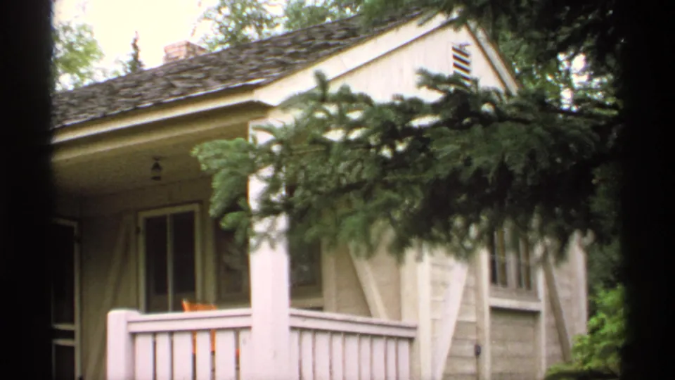1971 Isle Royale 8mm frame of a wooded cabin exterior, with evergreen branches crossing the view of a porch, windows, siding, and roofline.