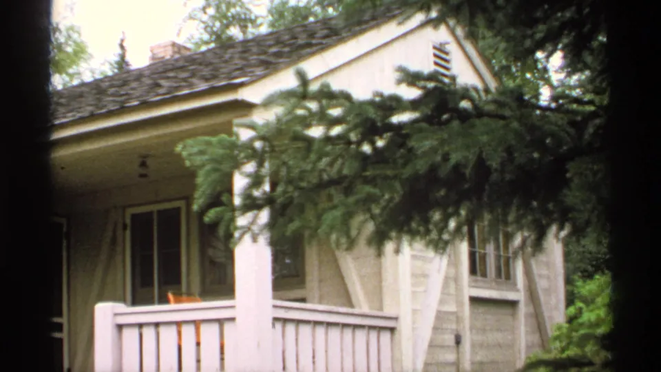 1971 Isle Royale home-movie frame showing a light-colored house exterior with porch railing, windows, roofline, and evergreen branches in front.