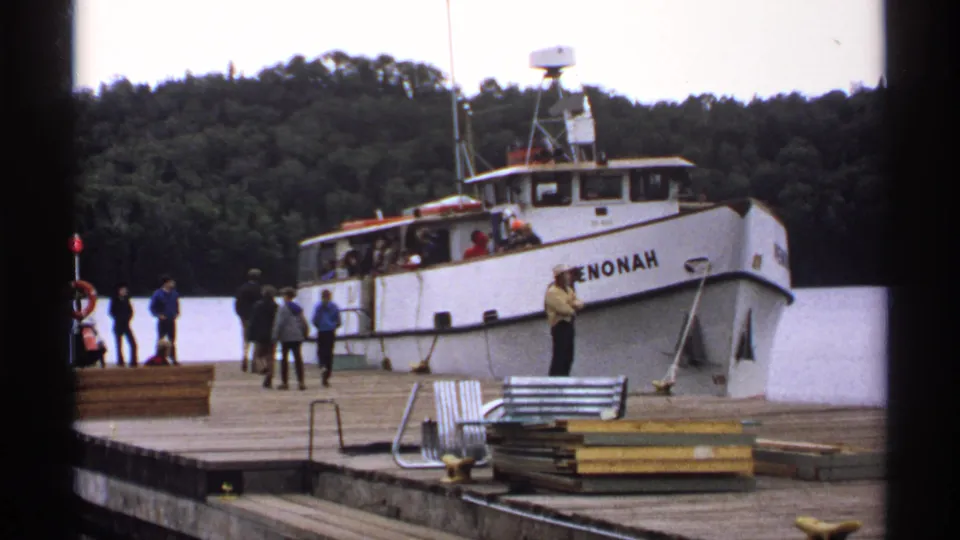 1971 Isle Royale frame with visitors on the wooden pier beside the PENONAH, with benches, stacked materials, and wooded shoreline visible.