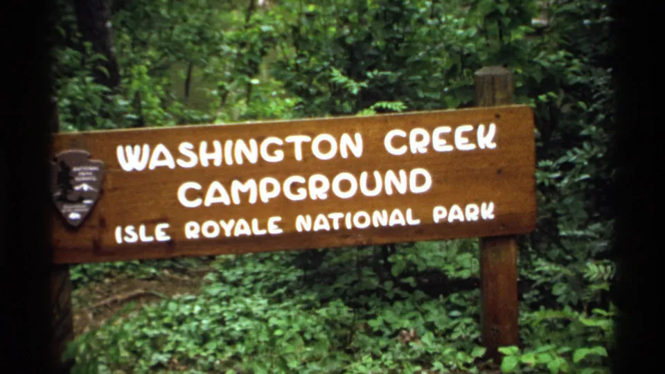 Close 1971 view of the Washington Creek Campground sign with Isle Royale National Park lettering and forest behind it.