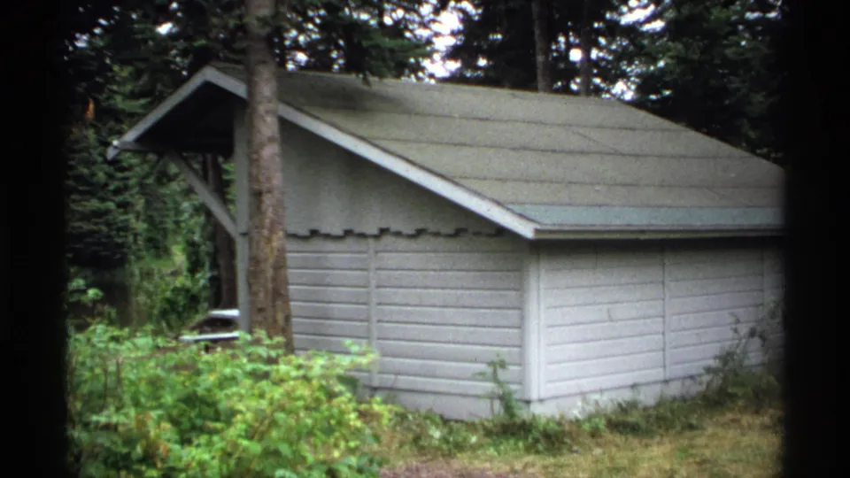 1971 Isle Royale footage frame holding on the same pale wooden forest structure with horizontal siding and surrounding evergreens.