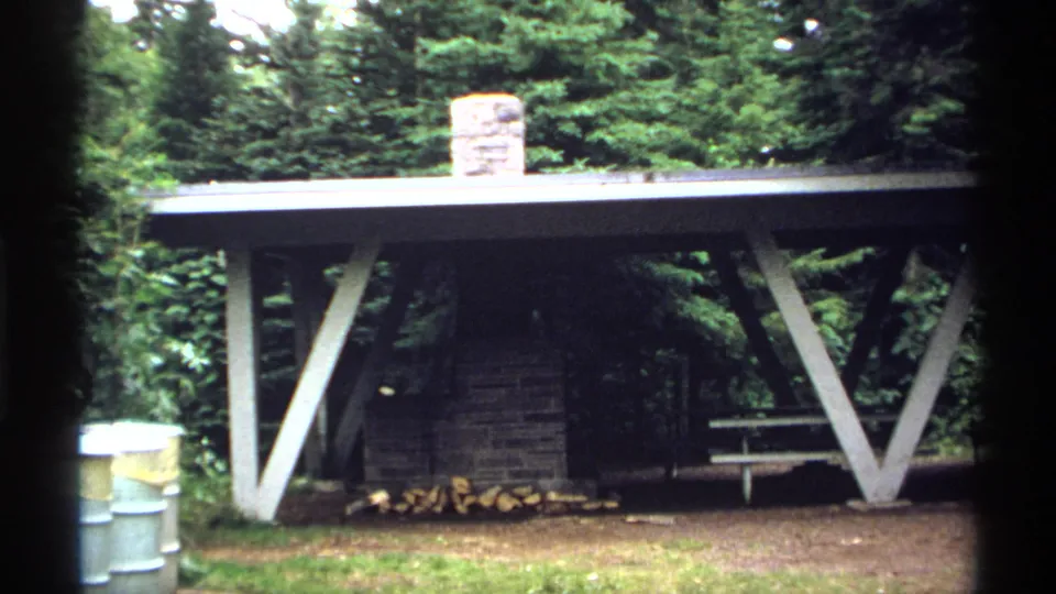 1971 Isle Royale home-movie frame showing an open rustic shelter with a stone fireplace, picnic table, stacked logs, and surrounding trees.