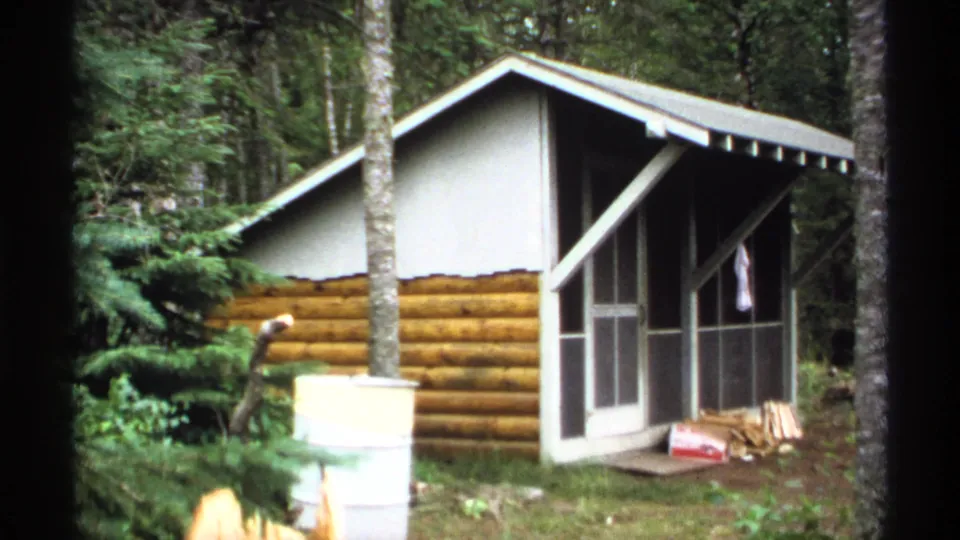 1971 Isle Royale frame of a rustic log-sided cabin with a screened porch, white upper gable, barrel, trees, and stacked firewood.