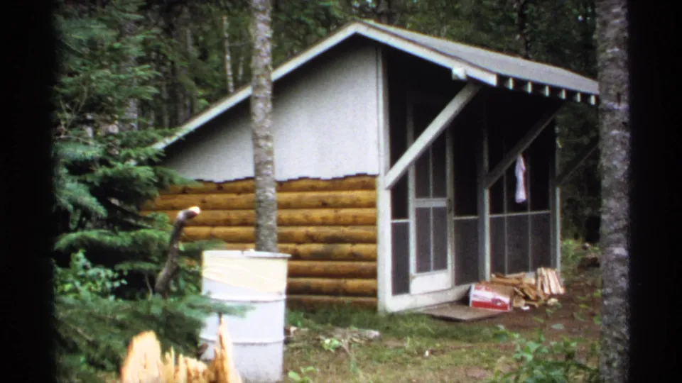 1971 Isle Royale 8mm still showing a small cabin in forest cover, with screened porch panels, a barrel, firewood, and leafy undergrowth.