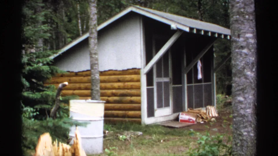 1971 Isle Royale frame focused on a log-sided forest cabin with screened porch, pale gable, metal barrel, stacked wood, and trees.