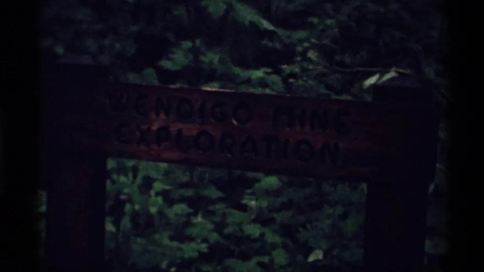 Wooden Wendigo Mine Exploration sign in the Isle Royale woods, seen in dim light with surrounding leaves and branches.
