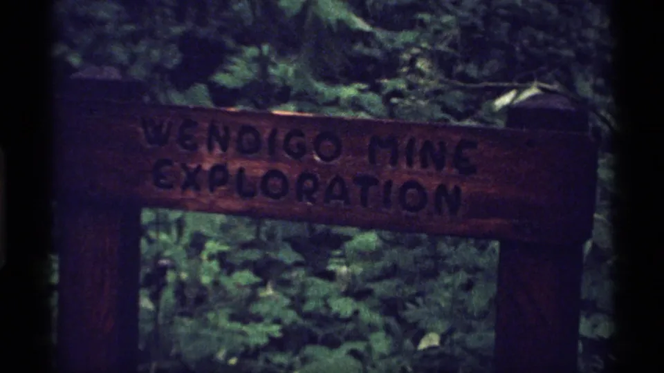 1971 Isle Royale home-movie frame with the Wendigo Mine Exploration sign centered against a dim wooded backdrop.