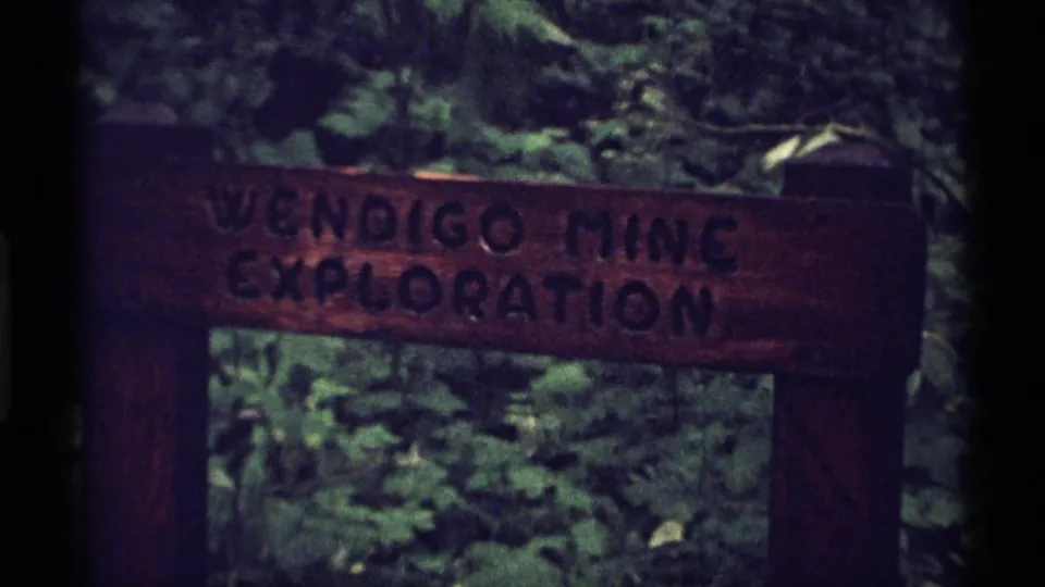 1971 Isle Royale 8mm frame showing the Wendigo Mine Exploration sign closer in the frame, with leaves and deep shade behind it.