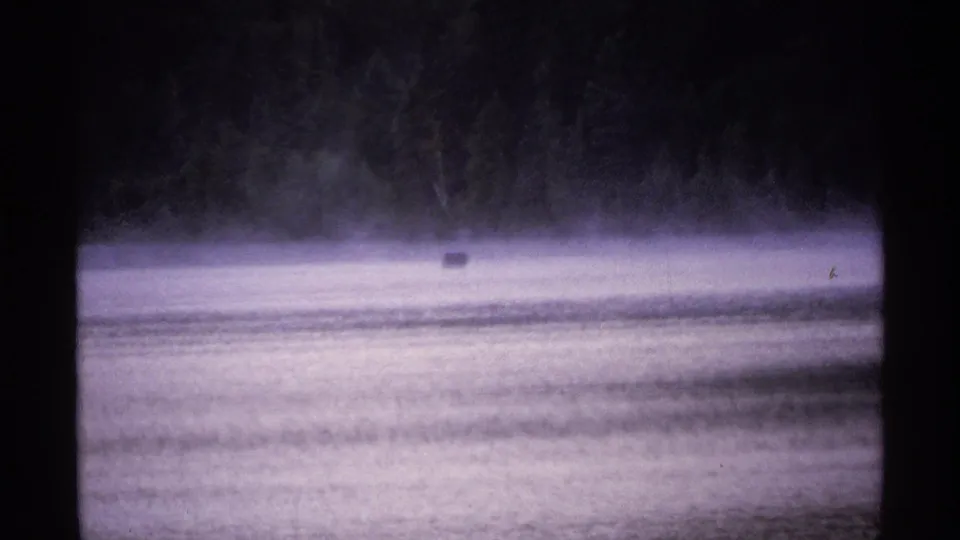 1971 Isle Royale 8mm frame with a small dark object on foggy lake water and a shadowed tree line in the distance.