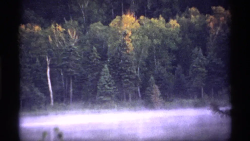 1971 Isle Royale home-movie frame showing a wooded shoreline through soft grain and mist over water.