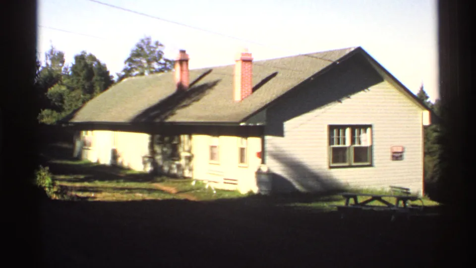 A modest wooden house on Isle Royale appears in bright sun, with a gray roof, red brick chimneys, windows, and trees behind it.