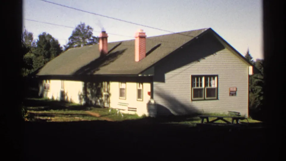 A steady 8mm frame of the Isle Royale house shows the shaded side wall, front windows, two chimneys, trees, and a picnic table at lower right.