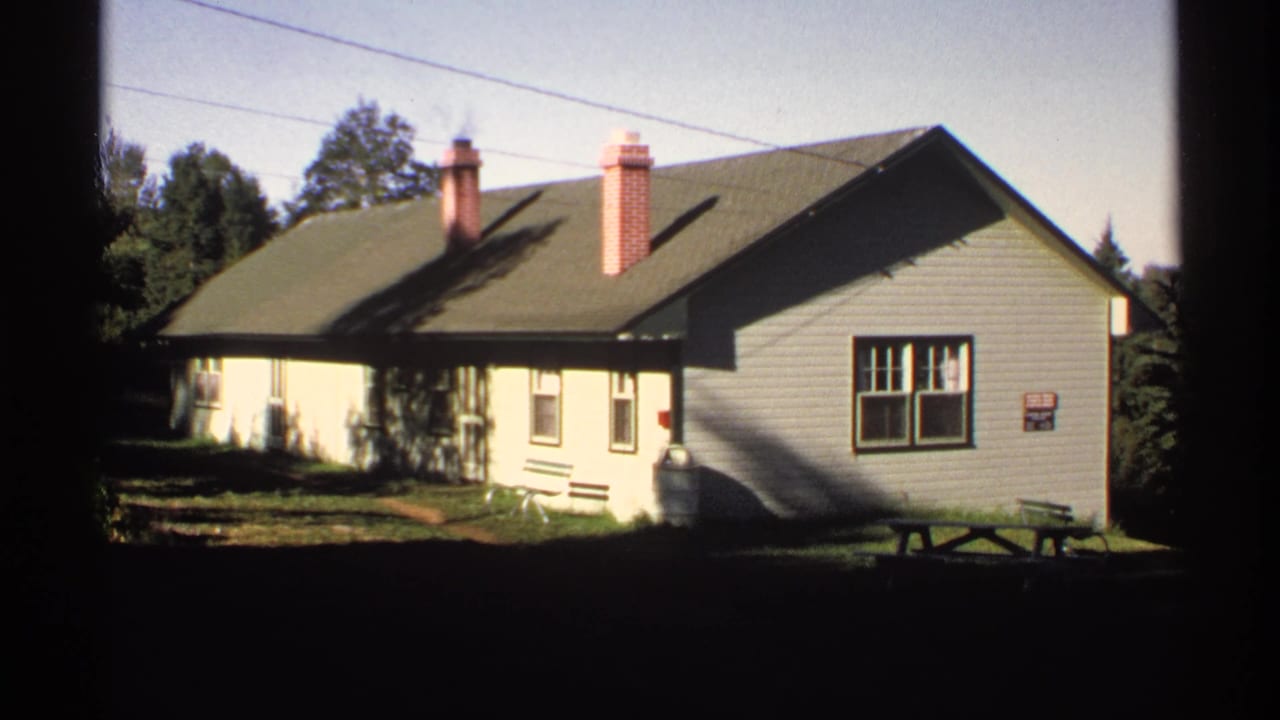 1971 Isle Royale lodge exterior with white siding, green roof, brick chimneys, surrounding trees, and a bright sky.
