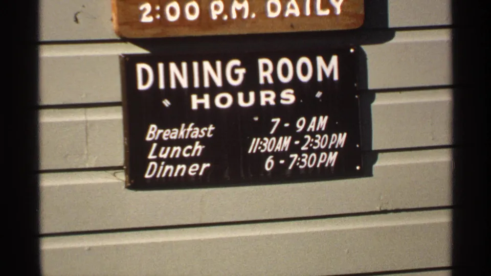 Close-up of a black dining room hours sign listing breakfast, lunch, and dinner times on the lodge exterior.