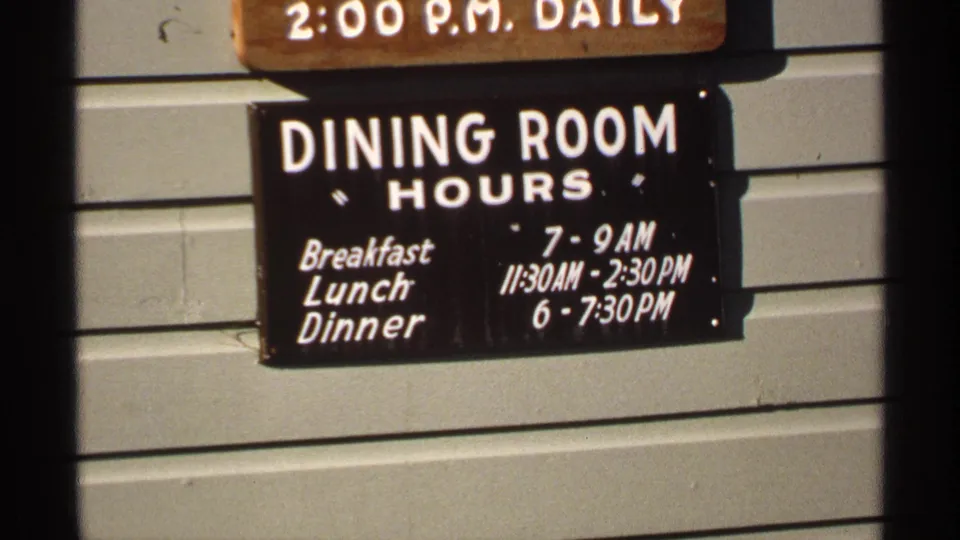 1971 Isle Royale dining room hours sign, with meal times painted in white on a dark board attached to exterior siding.