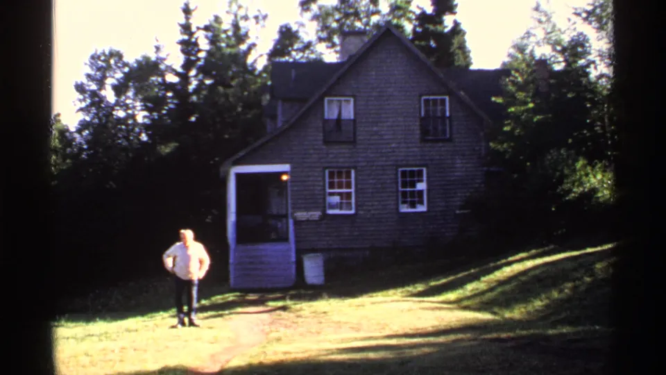 Wide 1971 Isle Royale home-movie view of a rustic two-story lodge with an adult man in a light shirt on the grassy lawn.