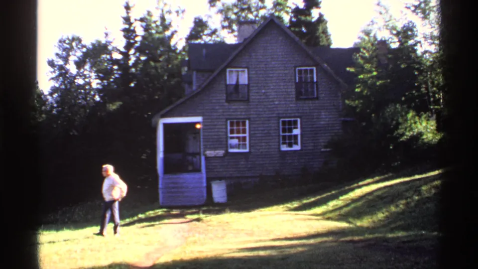 1971 Isle Royale 8mm frame showing an adult man walking across the lawn before a rustic shingled lodge surrounded by trees.
