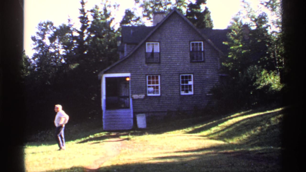 Man walking across grass in front of a rustic shingled house.