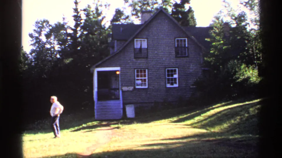 1971 Isle Royale home-movie frame showing a man crossing a sunny grassy yard before a shingled house with a screened porch.
