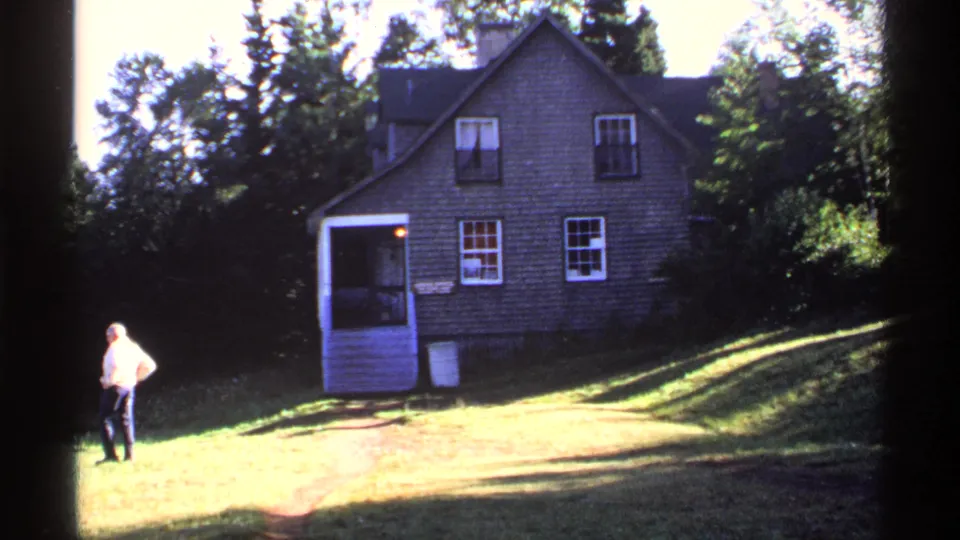 1971 Isle Royale frame with a man in a light shirt on the lawn near a rustic house and porch in bright sunlight.