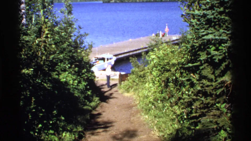 1971 Isle Royale footage frames a dock and small boat beyond dense green shoreline vegetation and bright blue water.