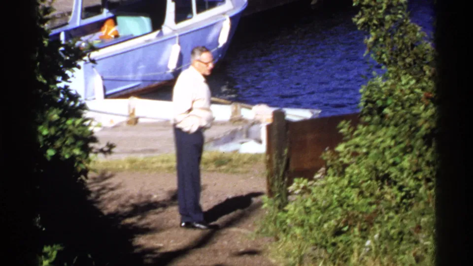 At Isle Royale in 1971, an adult man in a light shirt and dark trousers stands near a dock and moored blue boat.