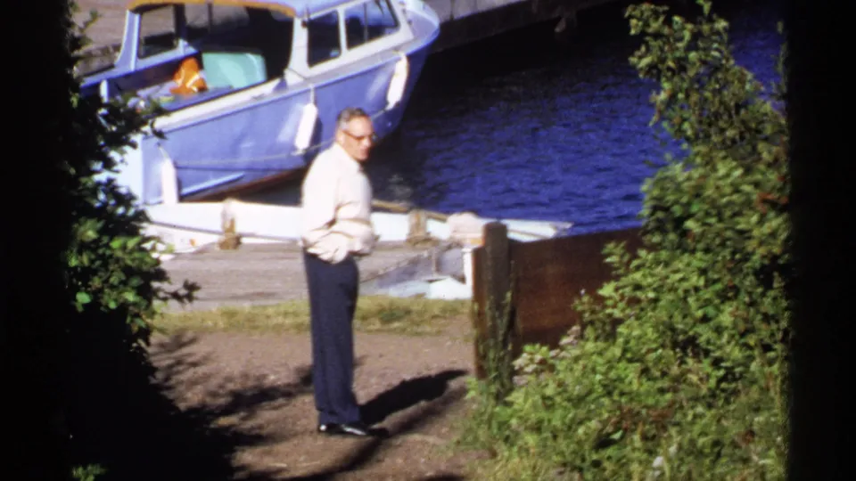1971 Isle Royale home-movie frame with an adult man looking toward the camera near a dock, blue water, and a moored boat.