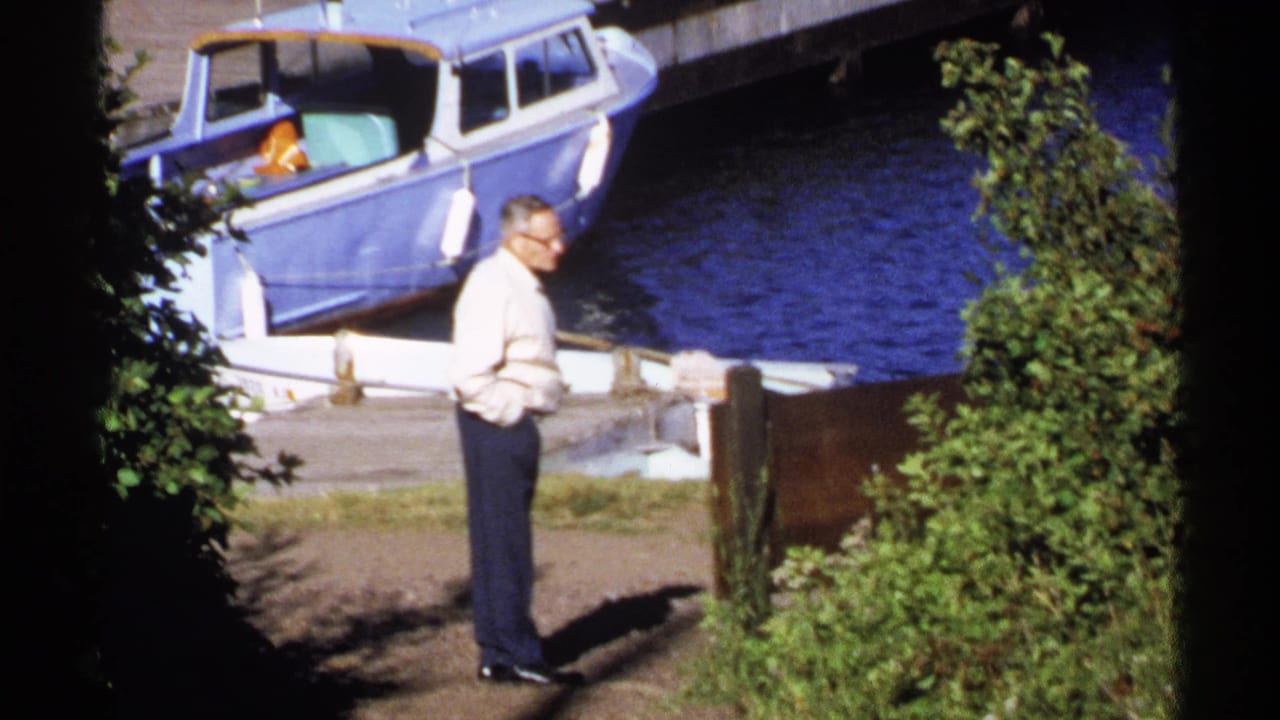 Older man standing on a dock beside a blue motorboat and water.