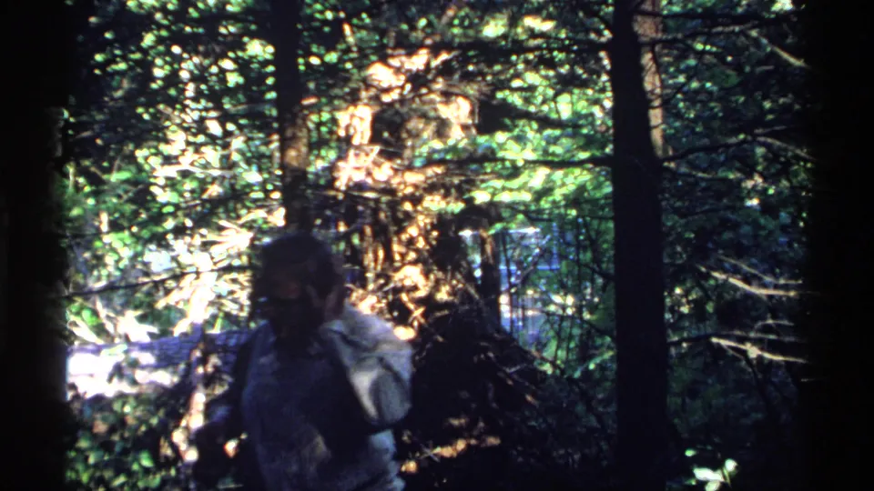 In a dark, leafy Isle Royale forest frame, the man appears partly shadowed with one hand raised near his face.