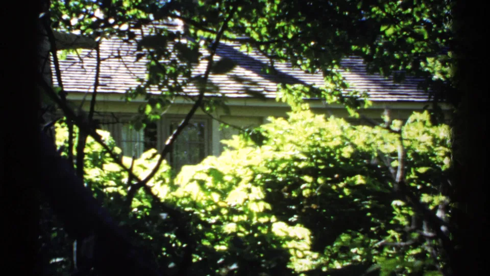 1971 Isle Royale home-movie frame with a pale house exterior partly visible through dense, sunlit vegetation.