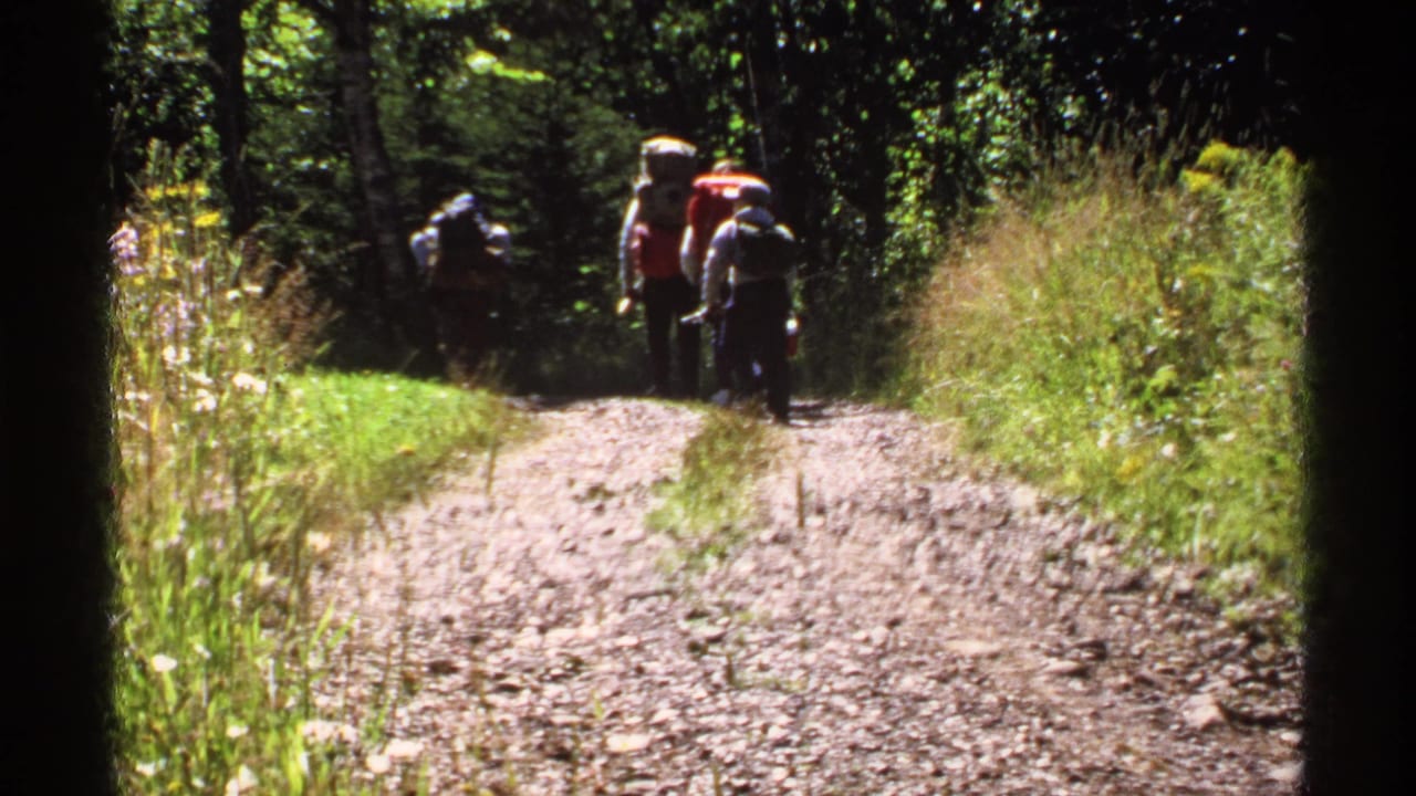 1971 Isle Royale home-movie frame showing hikers with large packs walking away along a sun-dappled forest path.
