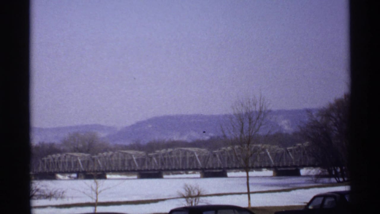 Wide winter river view with bridge, snow, bare trees, distant hills, and a parked car edge.