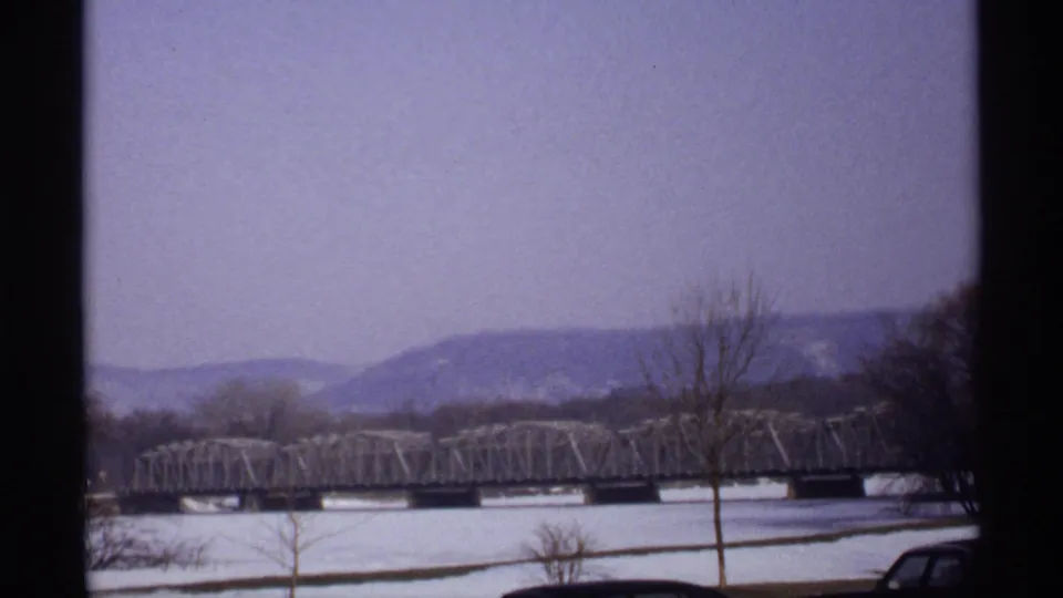 Soft 8mm frame of the 1971 La Crosse winter landscape, showing a metal truss bridge across a snow-covered river scene with bare trees and distant hills.