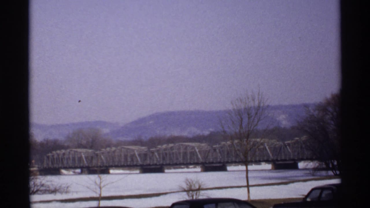 Snowy riverfront with a long truss bridge, bare trees, parked cars, and distant bluffs.