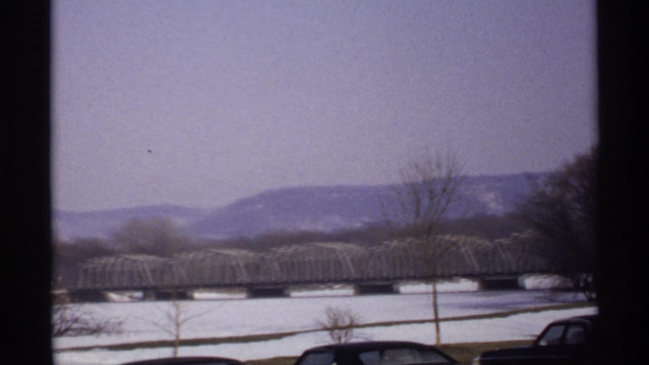 Truss bridge across a snowy riverfront with distant hills and leafless trees.