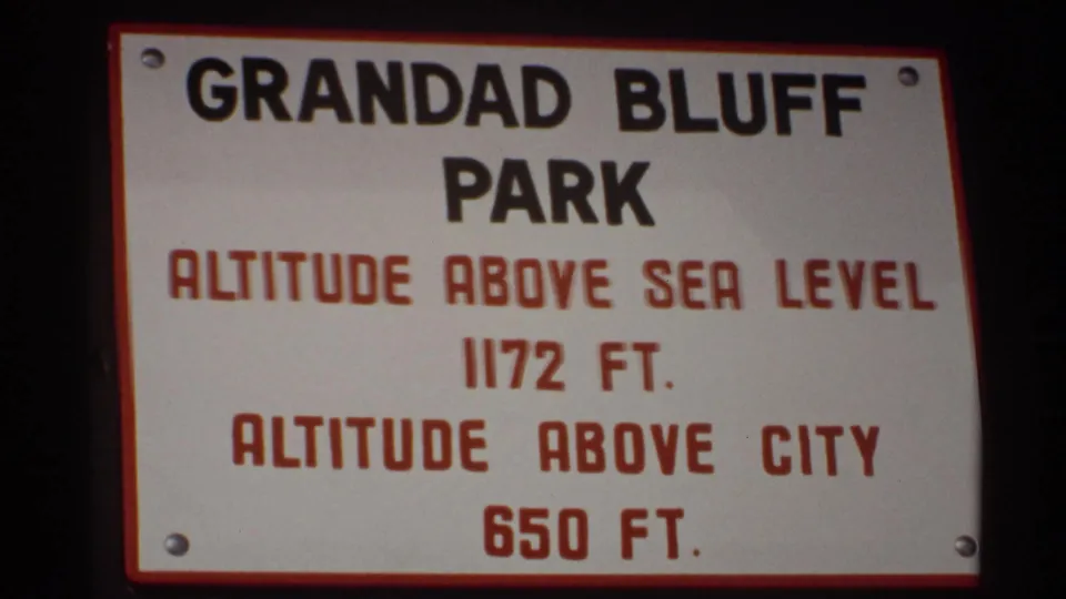 A 1971 La Crosse home-movie frame shows the Grandad Bluff Park sign with altitude details, with bridge and winter scenery behind glass.