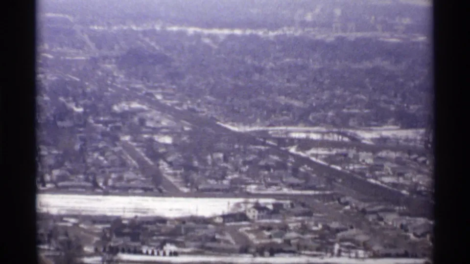 1971 8mm view across La Crosse from above, with roads, clustered buildings, snow-bright areas, and wooded hills beyond.