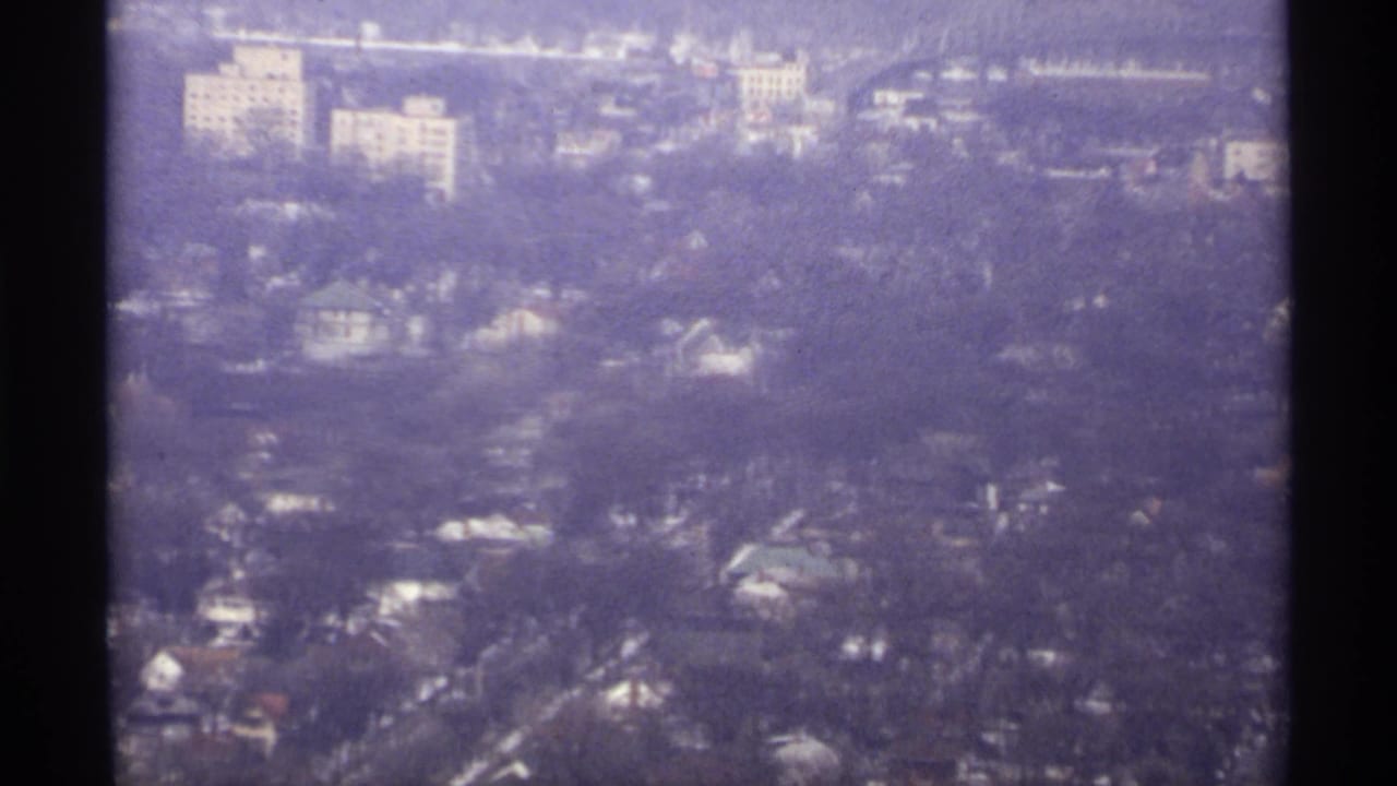 Aerial city view with houses, trees, roads, and larger buildings in the distance.