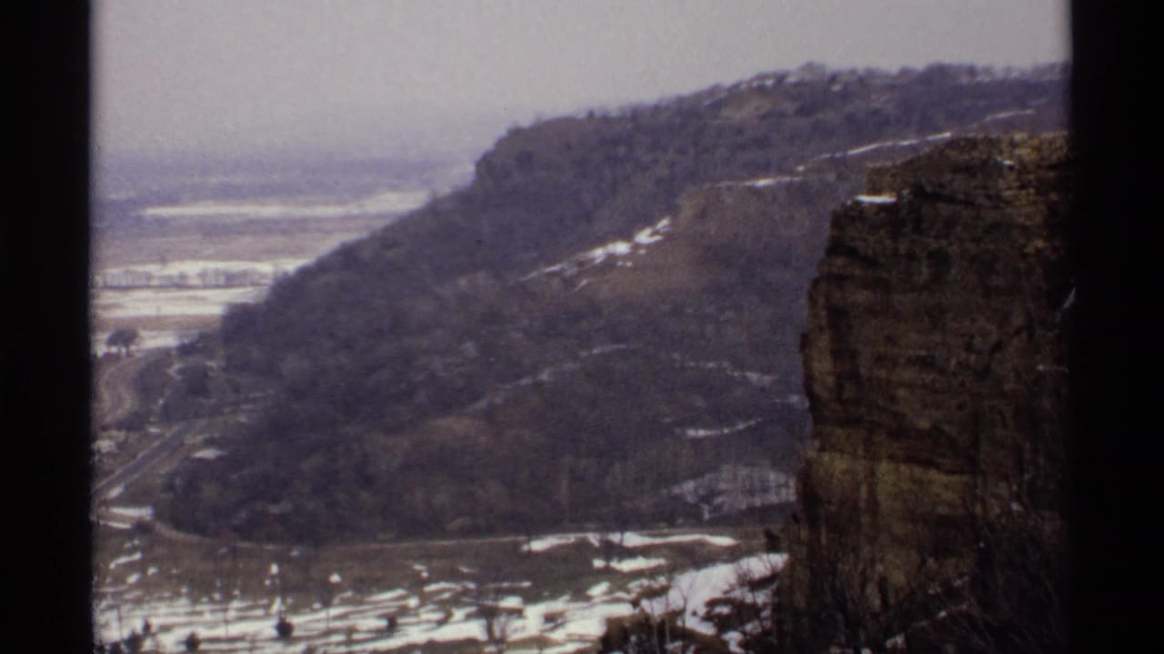 Wooded bluff and rocky cliff above a snowy valley with roads below.