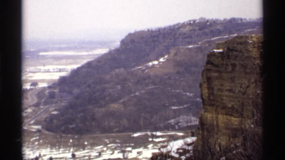 1971 8mm view of a La Crosse bluff slope, rocky outcrop, winter trees, curving road, and snow-covered valley floor.