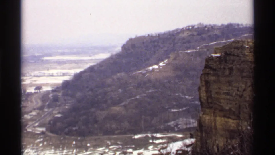 Soft 8mm frame of a La Crosse winter bluff, showing a rocky cliff edge, wooded ridge, snow patches, valley road, and pale sky.