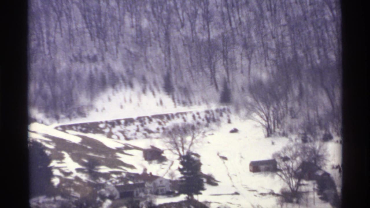 Snowy wooded hillside with a winding road and distant rural buildings.