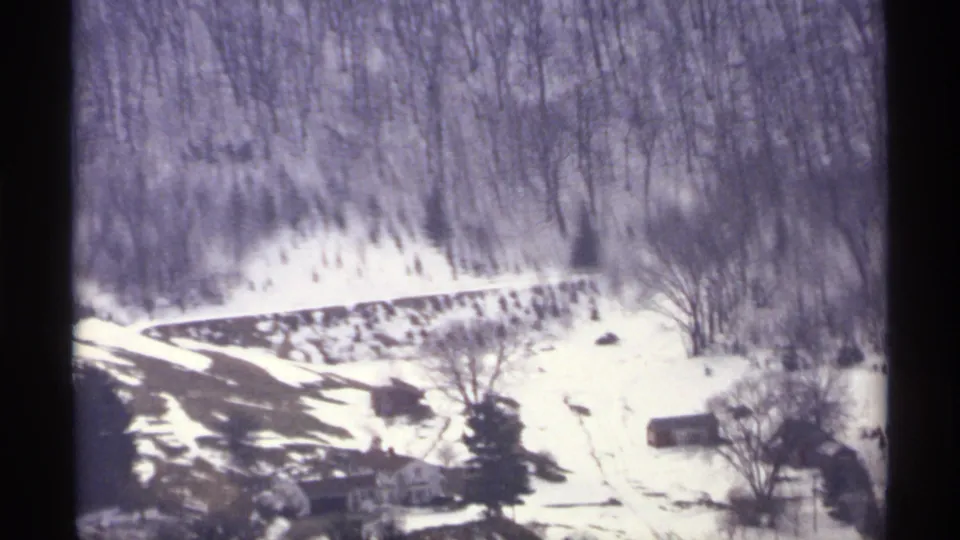 1971 8mm frame of a snowy hillside near La Crosse, Wisconsin, with bare woods, a curving road, and distant rural structures.