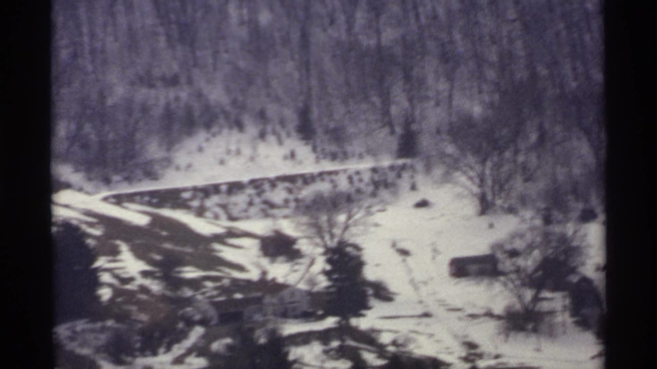 Snowy valley slope with wooded hillside, curving road, and small buildings.