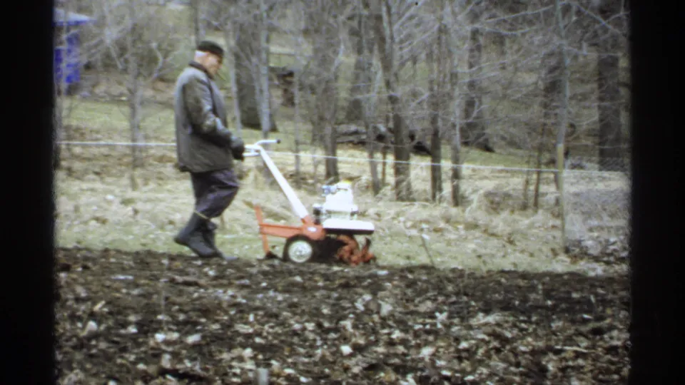 A man continues tilling soil with a small orange walk-behind machine in a rural La Crosse yard, with bare trees and fencing behind him.
